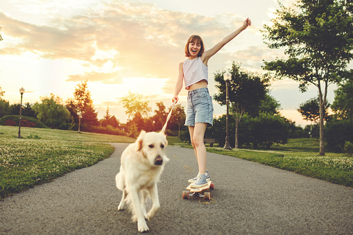 A Portrait of teenage girl petting golden retriever outside in sunset running on her longboard with dog