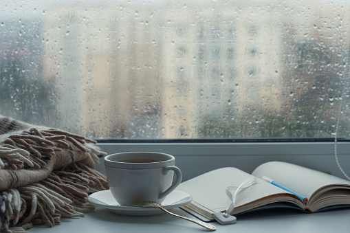 Rain outside the window. A cup of tea, a notebook with a pen and a blanket on the windowsill. Selective focus.