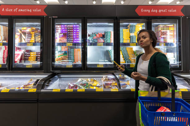 Browsing in The Frozen Section Mid-adult woman browsing in a refrigerated section while on a budget. She is looking for frozen food on a budget holding her mobile phone. She is living in the North East of England. smart phone technology lifestyles chain stock pictures, royalty-free photos & images