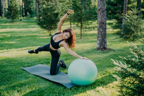 Flexible active woman in sportsclothes makes fitness exercises on karemat with fitball, raises arms and breathes fresh air in forest has morning workout poses over nature background. Healthy lifestyle la mujer activa flexible en ropa deportiva hace ejercicios de fitness en karemat con fitball, levanta los brazos y respira aire fresco en el bosque tiene poses de entrenamiento matutino sobre el fondo de la naturaleza. estilo de vida saludable