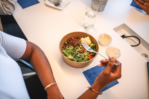 High angle view of healthy lunch bowl. Black mature woman is eating arugula and quinoa salad with dressing at office desk.