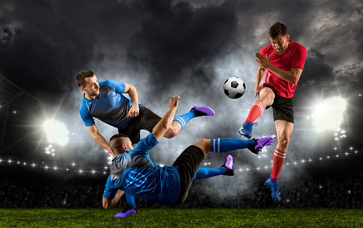 A male soccer player kicks a ball and soccer goalie jumping in motion for a ball while defending his gates on wide angle panoramic image of a outdoor soccer stadium or arena full of spectators under a sunny sky. The image has depth of field with the focus on the foreground part of the pitch. With intentional lensflares. Players are wearing unbranded soccer uniform.