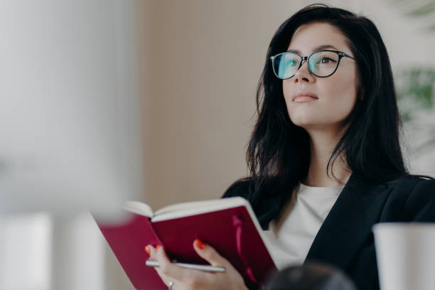 istock Beautiful woman with thoughtful expression, holds opened notepad, writes time management, makes plans for productive day, wears eyeglasses, poses indoor. Self employed female writes down ideas 1411238731