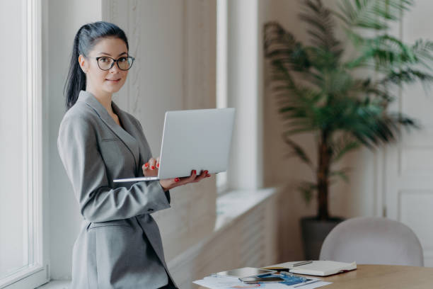 istock Woman administrative stands near desktop, holds modern laptop computer, uses wireless connection to internet, dressed in formal grey outfit, searches information on web page, works distantly 1411238752