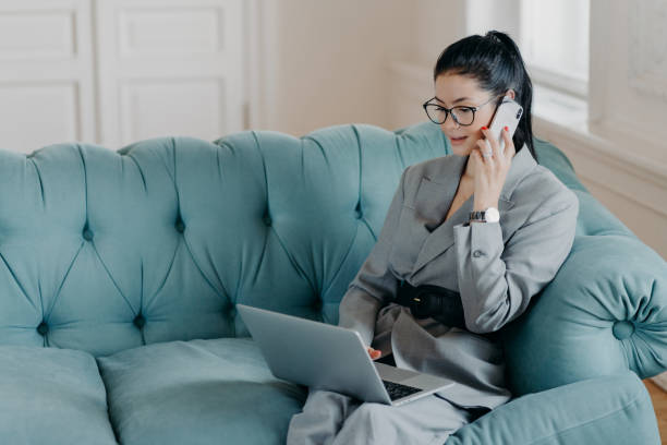 istock Busy female designer has cell phone conversation, sits in front of opened laptop computer on comfortable sofa, discusses new project. Businesswoman dressed in formal suit works remotely from home 1411238754