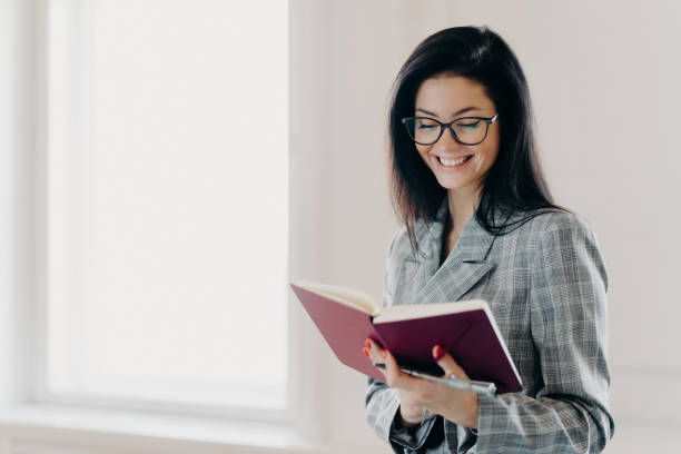 istock Satisfied brunette woman has happy expression writes down working plan in notepad has red manicure formal clothes records information for organization process wears spectacles notes financial schedule 1411238769
