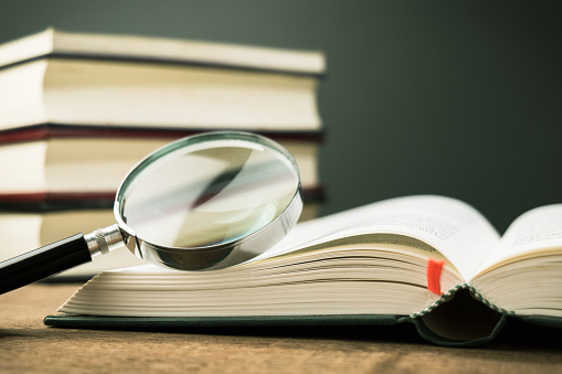 Magnifying glass on the opened book on the table, with a pile of books in background