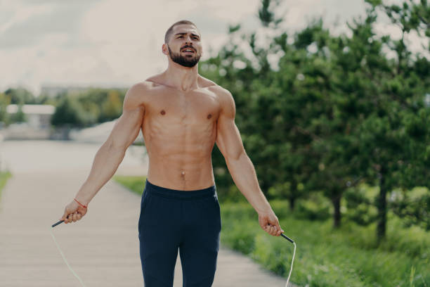 istock Athletic motivated man with naked torso concentrated on intense fitness training program jumps over skipping rope, poses in park has sporty body, breathes air deeply. Morning workout concept 1411243775