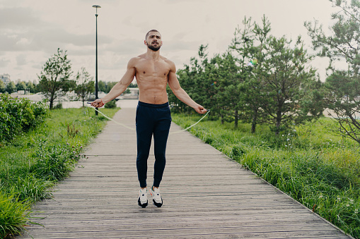 Sporty muscular man uses skipping rope for cardio training outdoor, has perfect body, breathes fresh air and demonstrates endurance, works out self development. Athlete guy with sport equipment