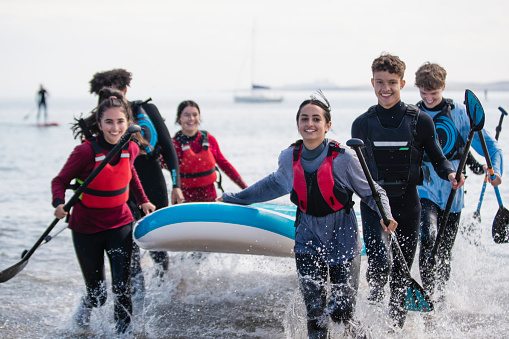 Group of mixed ethnic and age teens running in the ocean while carrying a multiple person paddle board together in the North East of England at Beadnell. They are wearing wet suits and life jackets to be safe and are laughing, having fun.