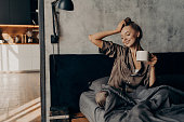 Portrait of young relaxed woman in stylish home suit waking up with cup of coffee in bed