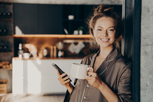 Lovely happy young woman having her morning coffee while checking new emails and notifications on smartphone standing in kitchen broadly smiling after waking up, spending weekend at home