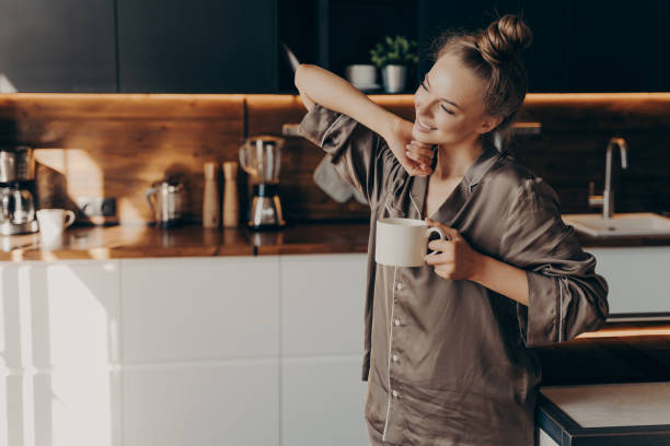 Young pretty relaxed woman in pajama with cup of coffee in morning Young pretty relaxed woman in comfy pajama stretching from sleep early in morning while holding cup of coffee in her hand, standing in stylish kitchen interior. Leisure time on average day at home pajamas stock pictures, royalty-free photos & images