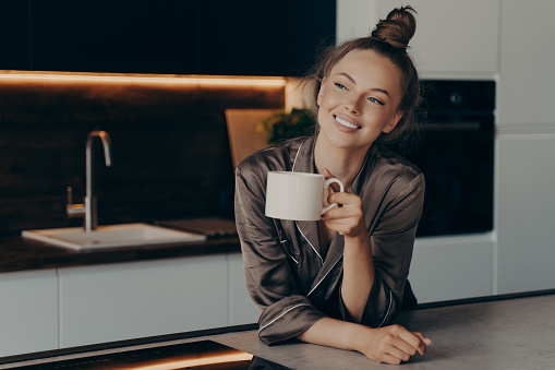 Happy smiling young woman in homewear enjoying aromatic morning coffee in modern kitchen after waking up before busy start of working day, positive female spending leisure time at home