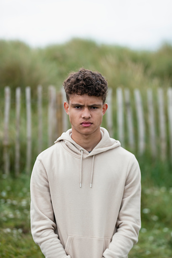 Medium waist up shot of a teenage boy looking at the camera with a serious expression. He is outdoors and is wearing a hoodie.