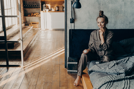 Young beautiful girl sitting on bed in pajama after waking up in stylish apartment bathed in morning sunlight, while holding and applying moisturizing face cream on her skin. Beauty treatment concept