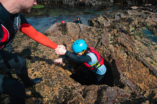 Group of mixed ethnic friends getting coasteering in the North East of England at Beadnell. They are wearing helmets and life jackets and one man is helping a friend climbing through a hole of a cave while they explore.