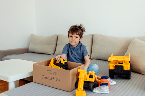 Photo of a Caucasian young boy, packing his old toys and some clothes into cardboard box for donation.
