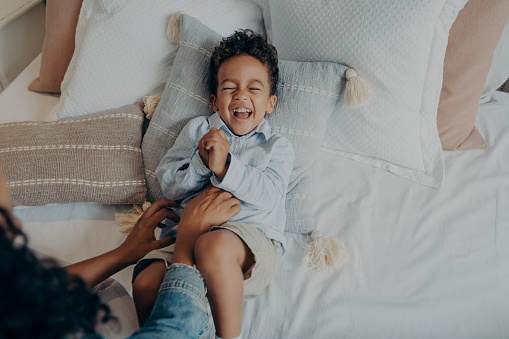 Woman mom tickles adorable mixed race little son with curly hair lying on pillows in bed. Cute baby boy in light colored casual clothes having fun and sincerely laughing. Family weekend leisure time