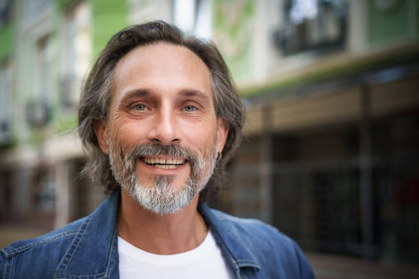 Happy handsome middle aged man standing outdoors on street of city looking at camera with a charming smile. Middle aged grey bearded man traveling in city smiling on camera wearing casual. feliz hombre guapo de mediana edad parado al aire libre en la calle de la ciudad mirando a la cámara con una sonrisa encantadora. hombre de barba gris de mediana edad que viaja por la ciudad sonriendo ante la cámara vistiendo ropa casual