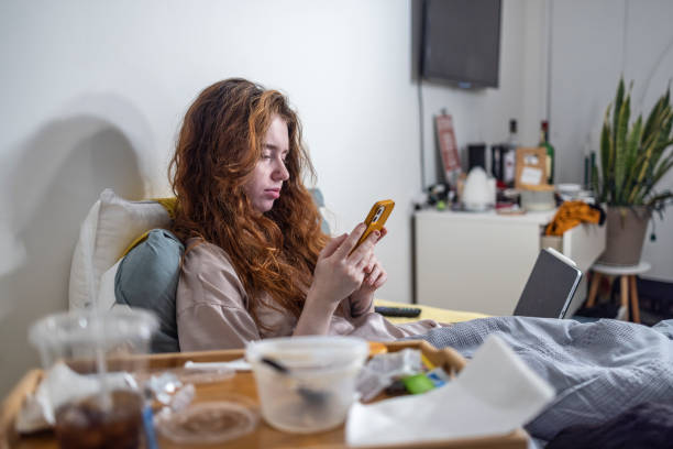 Internet and gadget addiction. Wasting time on smartphone. Woman lying in bed with phone and empty plastic containers. Focus on background with defocused foreground. stock photo