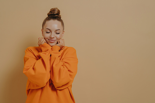 Happiness and excitement. Young delightful woman in orange sweatshirt smiling with closed eyes, feeling happy, isolated on beige background, keeping hands near face and dreaming of something good