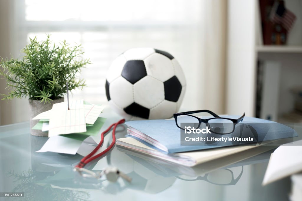Soccer ball on desk with books and whistle Soccer Ball on top of desk next to a stack of books with red whistle and papers next to plant, eyeglasses are on top of the stack of books plenty of room for a copy or background piece. Plant is also on desk and bookshelf and window in background. Book Stock Photo