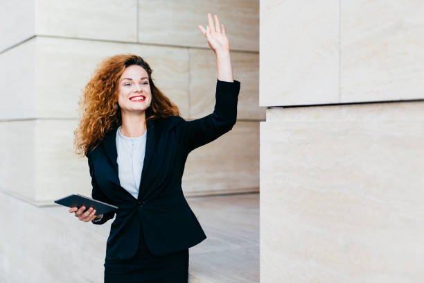 istock Smiling happy businesswoman dressed formally, holding in hands modern tablet computer, waving with her hand while noticing her companion, being glad to see him. Woman with curly hair saying `hello` 1411256643