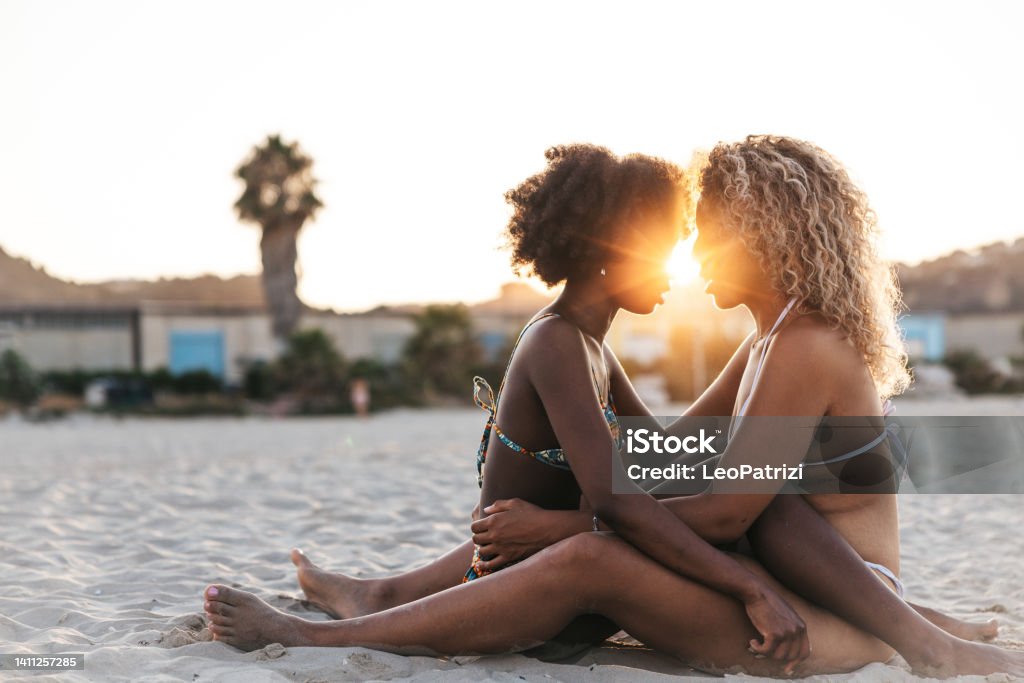 Two affectionate young women relaxing on the beach Cute and affectionate moments at sunset on the beach. On a vacation two friends embracing and kissing at the end of the day on the white sand. Gay Person Stock Photo