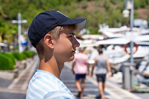 Handsome teenager guy 15-18 years old enjoying the view of marina.Teenage fashion. Close-up emotional portrait of a caucasian young man in a baseball cap. Sailboats anchor in a quiet bay.