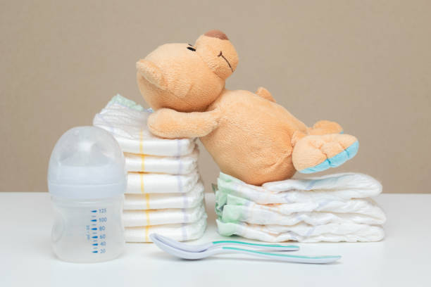 istock Stack of diapers with teddy bear toy lying on white table, bottle for milk and spoons. set for boy girl for baby shower, copy space 1411257415