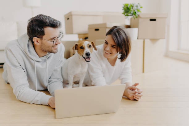 istock Lovely just married couple lie on floor, use laptop computer, creats design project for new house, their favourite dog poses between them, pile of cardboard boxes in background. Moving to new home 1411258463