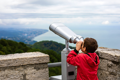 Rear view of a boy in a red windbreaker using binoculars on an observation deck on the coast of the sea and mountains.