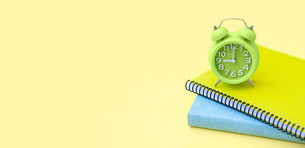 Green alarm clock, school accessories yellow and blue notebook, exercise book over pale background. Education, study, teenager concept. Back to school. Wide banner with copy space.