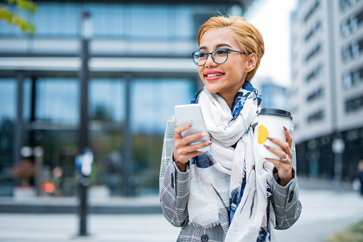 Young woman using mobile phone and holding cup of coffee in city
