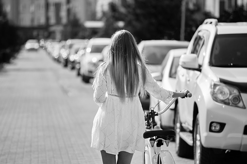 young blond woman in white dress walks away with bike near cars at city, rear view, monochrome
