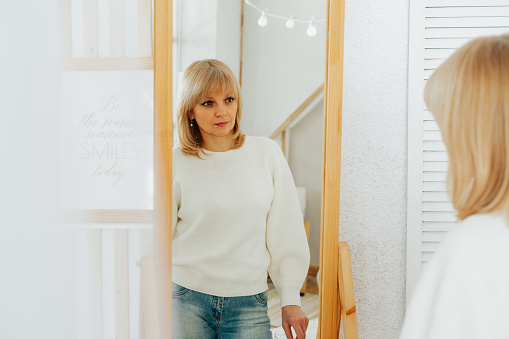 Beautiful smiling mature senior caucasian woman with blonde hair standing near mirror and looking at reflection at home.