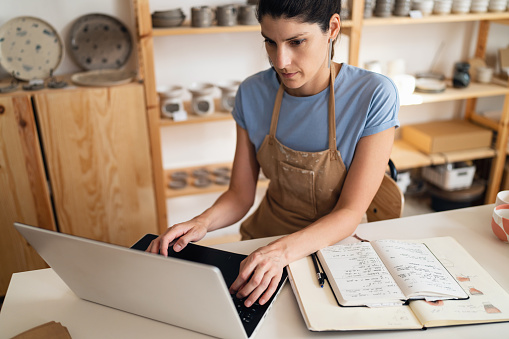 Young Caucasian female small business owner working on laptop from her modern pottery studio