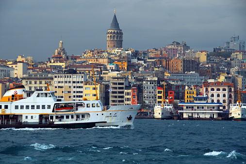 Istanbul, Turkey / Türkiye: skyline of Galata / Karaköy neighborhood in Beyoglu district seen from the Bosphorus strait, with passing ferry and the Galata Tower in the center. Karaköyis located at the mouth of the Golden Horn on the European side of the Bosphorus.