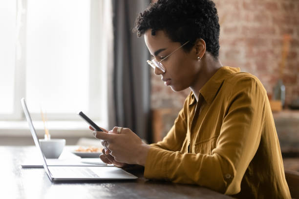 istock Serious African woman sit at desk with laptop using smartphone 1411275954