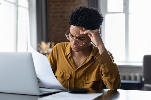 Young African woman read bad news frowning feels annoyed, frustrated due financial overspend, bank debt or bankruptcy, analyze financial report sit at desk looks troubled. Finances problems, concept