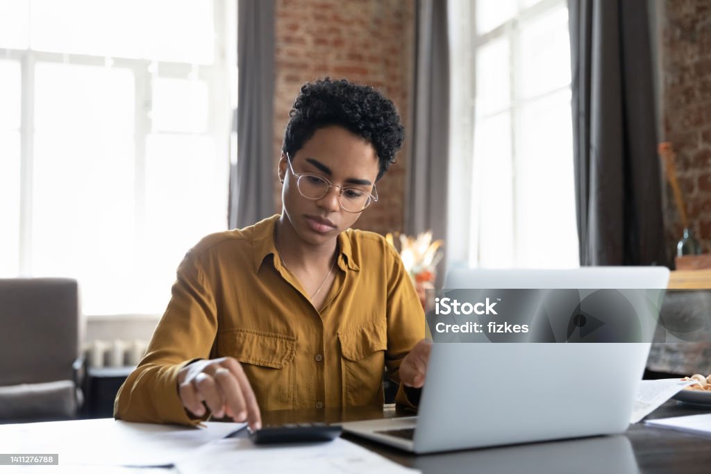 African woman sit at desk calculates bills do accountancy job Serious African woman sit at desk with laptop, analyzing bills, do accounting job, review utilities, calculates summary for payment through electronic bank app. Finance management, bookkeeping concept Bookkeeping Stock Photo