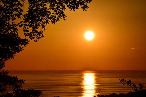 The sun is setting over the ocean.  The sky is orange and many clouds are making the sunlight very dramatic.  The ocean is very calm.  The image is very relaxing and shows the beauty of our planet.