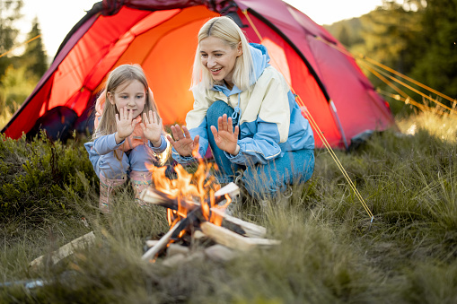 Young woman with her little girl sit by the campfire, bonding together while travel on nature. Mother with daughter warming up at campsite