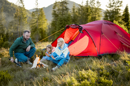 Young adult couple sit with a little girl by the campfire at campsite, traveling with tent on nature. Family spend summer time hiking in the mountains