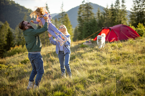 Young caucasian couple with little girl and dog have fun while travel with tent in the mountains. Happy family summer vacation at campsite. Father tossing daughter in his arms