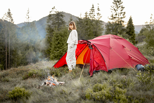 Young woman enjoys nature while standing at campfire near camping tent in the mountains. Concept of escaping to nature