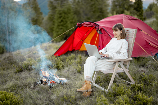 Young woman works on laptop while sitting relaxed on chair by the campfire, traveling with tent in the mountains. Concept of remote work and escape to nature