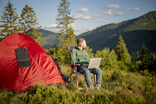 Man works on laptop while traveling with tent in the mountains. Charging computer with portable solar panel hanging on tent. Concept of remote work at campsite and renewable energy