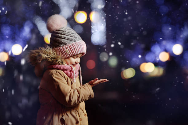 At Christmas, a child in a winter jacket and a hat with a pink pom-pom catches snowflakes at night against the background of the bokeh of New Year's garlands.
